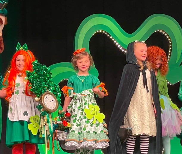 Little Girls Dressed as a wide Assortment of Leprechauns stand on a Stage in front of Lit Shamrock Leprechaun Contest