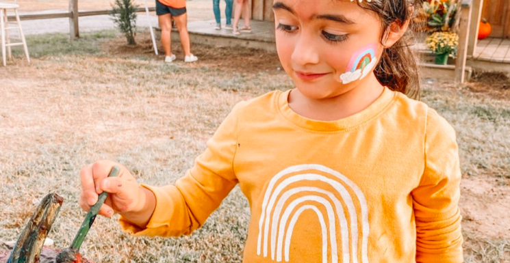 Little girl with face paint painting a pumpkin at a country pumpkin patch in Autumn
