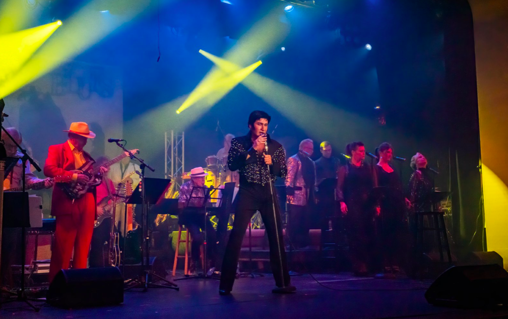 Elvis impersonator singing on stage with a full band behind him and yellow stage lights shining down